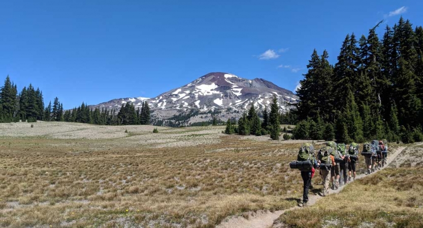A group of outward bound students carrying backpacks hike along a trail through an open field. There is mountain spattered with snow in the background. 